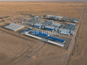 Aerial panorama of the 557-unit modular construction camp for the Saudi Arabia SDW project, showing single-storey prefabricated steel buildings arranged in functional zones across a desert site, with blue-roof office buildings, white accommodation blocks, internal roads and fenced perimeter