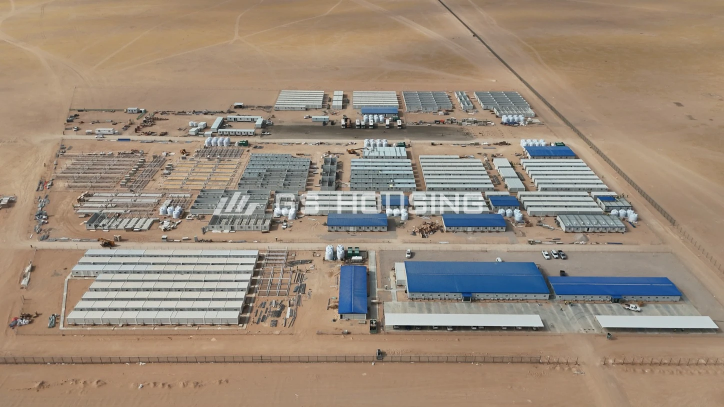 Aerial view of the SDW modular construction camp in Saudi Arabia, showing separate functional zones including blue-roof office buildings, rows of white prefabricated steel accommodation blocks, internal access roads and shared facilities within a fenced desert compound