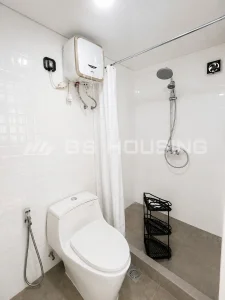 Completed private bathroom interior within a modular dormitory unit at the Indonesia workforce accommodation project, showing a toilet, shower with rainfall showerhead, wall-mounted water heater, exhaust fan, tiled walls and grey floor tiles