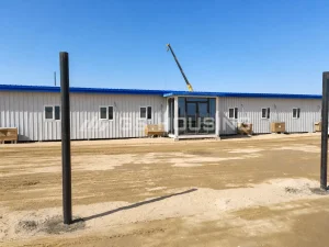 Exterior view of a blue-roof prefabricated steel building at the SDW modular construction camp in Saudi Arabia, showing white corrugated metal wall panels, a glass entrance porch, windows and level footings on a sandy desert site
