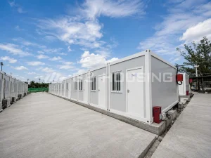 Ground‑level view between rows of white modular construction site accommodation units at the Bahamas stadium renovation project, showing individual doors, barred windows, air conditioning units, concrete footings and paved access routes