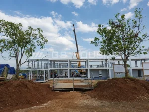 Construction site of the 132-unit Indonesia modular dormitory project showing steel frame structure with crane lifting prefabricated modules, workers on site and trees preserved around the construction area