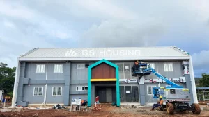 Front elevation of a two-storey modular dormitory building under construction at the Indonesia workforce accommodation project, showing grey exterior walls, white roof, decorative teal and brown entrance portal, and workers on a boom lift completing exterior installation