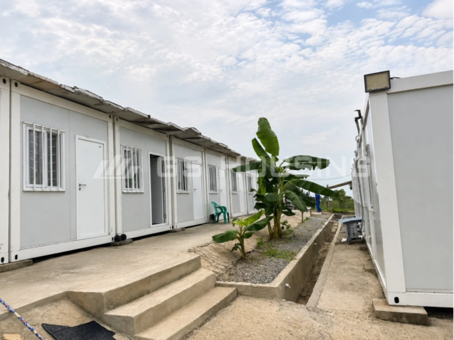 Walkway between rows of white flat pack container accommodation units at the Arua Enyau water supply project in Uganda, with concrete access steps, drainage channels and tropical banana plants