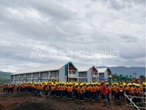 Large group of workers in orange uniforms gathered in front of completed two-storey modular dormitory buildings at the 132-unit Indonesia workforce accommodation project, with mountains in the background