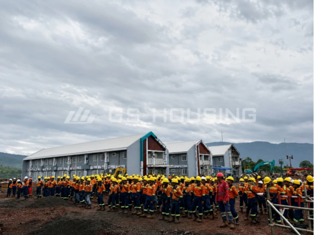 Large group of workers in orange uniforms gathered in front of completed two-storey modular dormitory buildings at the 132-unit Indonesia workforce accommodation project, with mountains in the background