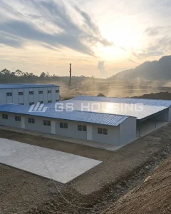 View of the FHT nickel smelting project construction site accommodation, showing single-storey blue-roof prefabricated steel buildings with a covered walkway, with the active industrial site, drilling equipment, dust and mountainous terrain visible in the background
