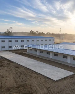 Exterior view of prefabricated steel buildings at the FHT nickel smelting project, showing a single-storey building with multiple doors and barred windows in the foreground and a two-storey building behind, with concrete access roads, drilling equipment and dust from the active industrial site in the background