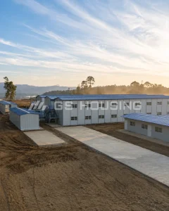  Overall view of the construction site accommodation for the Indonesia FHT nickel smelting RKEF project, showing blue-roof prefabricated steel buildings including a two-storey block with external stairs and single-storey blocks, with concrete access roads and a mountainous forested backdrop