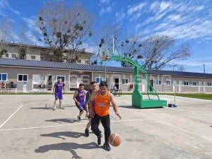 Single-storey workforce accommodation buildings at the Chile metro construction project, with a basketball court in the foreground where workers are playing, rows of doors and windows along the building, and trees in the background