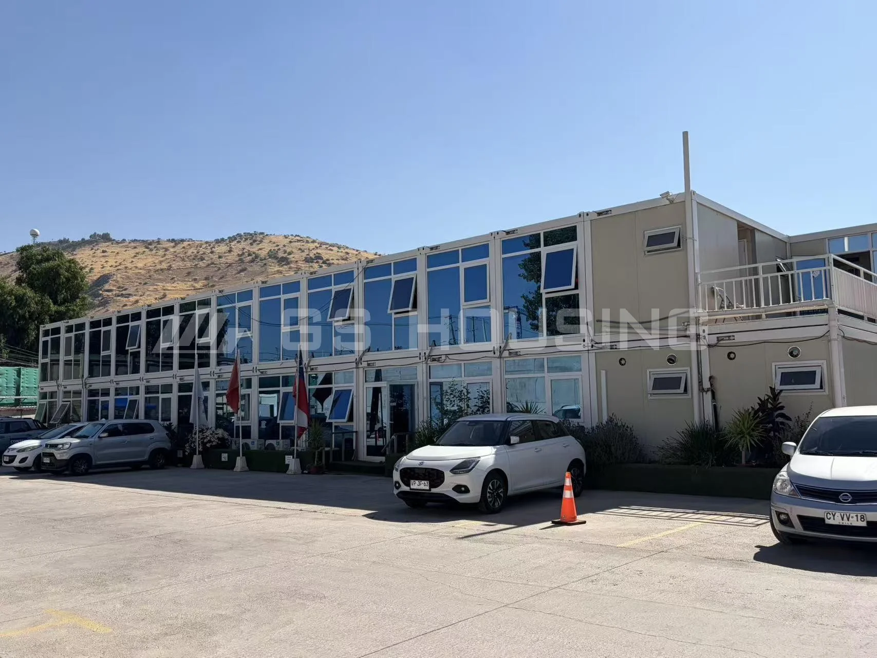 Two-storey modular site office building at the Chile metro construction project, with blue-tinted upper-floor windows, a balcony, parking area and dry hills in the background